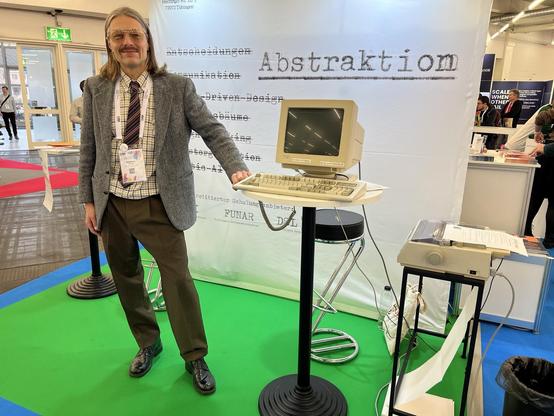 Photo of Active Group’s trade-show booth, with Marcus Crestani in front. The back fall features the word “Abstraktion”. The foreground contains a CRT terminal and a dot-matrix printer.