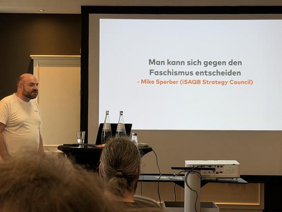 Foto von Christoph Iserlohn vor Folie, auf der Steht “Man kann sich gegen den Faschismus entscheiden —Mike Sperber (iSAQB Strategy Council)”.