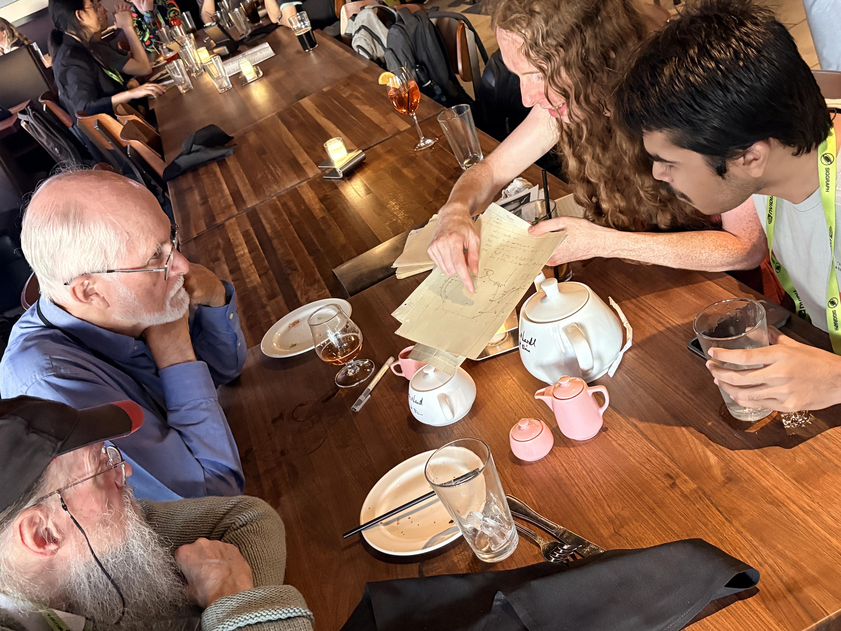 Martin Newell, Jim Blinn, Thalia Archibald, and Kai Deshpande at the 1976–2026 Utah reception at the Yard House, LA Live. I am showing my copy of Martin's original drawing of the Utah teapot. On the table are my signed Melitta teapot, 3D printed Utah teapot, and Melitta children's pink coffee set.